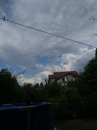 A low-angle shot shows a partly cloudy sky above a partially obscured house and surrounding greenery. The house has a brown-tiled roof and several windows, some of which are open. A chimney pipe is visible protruding from the roof. Dense foliage with some orange flowers surrounds the house. In the foreground, a dark blue inflatable pool with white supports is visible. A person is partially visible inside the pool, looking down. A black cable runs diagonally across the frame from the upper left to the lower right. The scene appears to be set outdoors, possibly in a residential area, given the house and pool. The time of day is indeterminate due to the cloud cover, but the lighting suggests daytime. The weather appears to be mixed, with both cloudy and clear sky visible. No visible text is present.