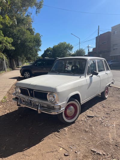 A white vintage sedan, possibly a Renault 6, is parked on a dirt and gravel shoulder on the right side of a paved street in Quilmes Oeste, Argentina, under a clear blue sky. The vehicle features chrome bumpers, round headlights, and red-centered wheels with white-walled tires. Its front license plate displays "USF 620" and "ARGENTINA." Strong sunlight casts distinct shadows to the left and rear of the car.
To the immediate left, a dark-colored Jeep Renegade SUV is parked partially on a concrete sidewalk that runs parallel to the street. Further left, a row of mature trees with dense green foliage lines the sidewalk, creating additional shadows.
On the right side of the street, beyond the paved section, several buildings are visible. These include a two-story structure with exposed red brick and small rectangular windows, and an adjacent multi-story light-colored building with multiple windows. Overhead, a network of utility wires spans between multiple utility poles and a streetlamp. No individuals are visible in the scene.