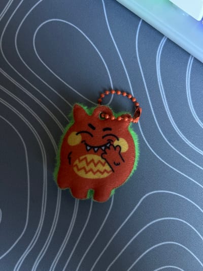 This image depicts a plush keychain, shaped like a cartoonish, orange-red monster with fluorescent green fuzzy edges, resting on a dark grey desk mat. The monster features a smiling expression with visible white fangs, yellow-orange circular cheek markings, and a yellow-orange zigzag pattern on its lower torso. One of its hands is raised, with fingers touching its mouth. An orange plastic beaded chain is attached to a fabric loop at the top of the monster's head. The desk mat is patterned with numerous thin, white, wavy, parallel lines. A partial view of a white electronic keyboard with illuminated keys is visible in the upper right corner of the frame. The scene is situated indoors, likely on a desk surface, in the city of Enem, Russia.