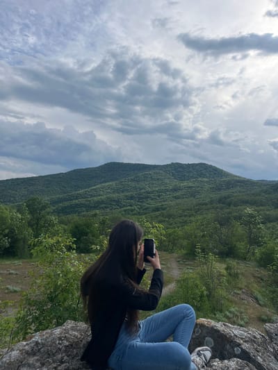 The image depicts a landscape near Kamchia, Bulgaria. A person is sitting on a rock overlooking a forested hillside and cloudy sky. They are wearing jeans and a black jacket, and are holding a mobile phone, possibly taking a photo. A portion of a second person's legs is visible next to them on the rock. The environment is lush with greenery, suggesting a forested or mountainous area.