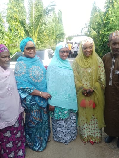 The image displays an outdoor scene in Unguwa Uku Kauyen Alu, Nigeria, featuring five individuals standing in a line, facing the viewer.
From left to right:
1. An elderly woman, partially visible, wears a pink head covering and a patterned purple and white dress. She looks towards the left.
2. A middle-aged woman with dark sunglasses wears a turquoise headwrap and an embroidered turquoise shawl over a patterned blue dress. She smiles directly at the camera, displaying gold bracelets and rings.
3. A middle-aged woman with glasses wears a light blue hijab and a matching embroidered shawl over a green and white patterned top and grey patterned skirt. Her hands are adorned with henna, and she looks towards the right.
4. A young woman wears a cream and gold patterned head covering, a sheer lime green veil, and an embroidered light green dress with red shoes. Her hands bear intricate henna designs, and she holds a small red handbag while smiling at the camera.
5. On the far right, an elderly man in a brown traditional cap and matching boubou-style garment, with a grey beard, looks directly forward.
The background consists of dense green foliage, including tall palm-like trees. A white vehicle is partially visible behind the second woman. Indistinct figures and structures are visible in the distance under bright natural light, indicating an outdoor gathering. The ground beneath the individuals is unpaved, appearing to be light brown earth or gravel.