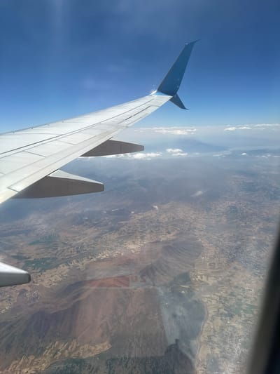 This is an aerial photograph taken from an airplane window, looking down at a landscape. The dominant feature in the foreground is the wing of the aircraft, painted white with a blue wingtip. The wing extends from the left side of the image across the upper portion. Below the wing, a vast, arid landscape unfolds, characterized by dry, brown earth, interspersed with fields of varying shades of green and tan, suggesting agricultural activity. Rolling hills and mountains are visible in the distance, some shrouded in haze. In the far distance, a body of water, likely a lake or sea, is visible, with a distant landmass or mountain range on the horizon. The sky is a clear, bright blue with a few wispy white clouds. The image appears to be taken during daylight hours, with the sun casting light and shadows on the landscape. The specific location in Şarkikaraağaç, Turkey, cannot be definitively identified from the visual information alone without external data.