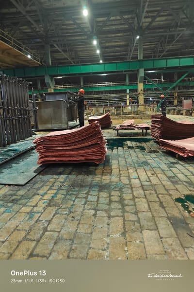 The image depicts an industrial interior, likely a copper smelting or processing plant in Jezqazğan, Kazakhstan. In the foreground, large stacks of reddish-brown copper sheets are laid out on a wet, cobbled floor. The floor itself is marked with blue and yellow stains, indicating potential chemical spills or residue.
Two workers, wearing orange hard hats and dark clothing, are visible in the middle ground. One worker is standing near a large, metal vat or tank, appearing to be engaged in an activity related to the copper sheets. Another worker is seen further back, also with an orange hard hat, near some machinery.
The environment is characteristic of heavy industry, with high ceilings, exposed structural beams, and industrial lighting fixtures illuminating the space. The overall atmosphere suggests ongoing industrial operations. The image is overlaid with a watermark indicating "OnePlus 13" and camera settings: "23mm f/1.6 1/33s ISO1250", along with a photographer's signature.