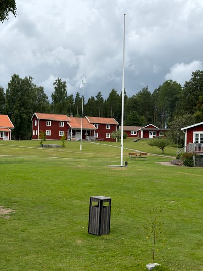 The image depicts a spacious, well-maintained grassy area with several red-painted wooden buildings that appear to be cottages or cabins, each with orange tile roofs and white-trimmed windows. In the foreground, a dark grey wooden trash receptacle stands on the lawn. Further into the scene, a tall, slender flagpole is prominently positioned. Behind the buildings, a dense forest of green trees forms a backdrop against a cloudy, overcast sky. Scattered across the lawn are a few small trees and some park benches. The environment suggests a peaceful, possibly recreational, setting, likely a campground or resort, in the municipality of Aneby, Sweden, given the context. There are no people or animals visible in the image.