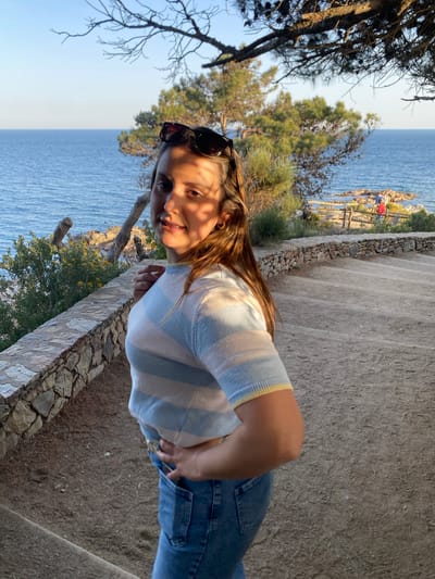The image is a low-angle, rotated portrait of a woman on a stone staircase near the coast. The woman has long brown hair and is wearing sunglasses, a striped top, and blue jeans. She is smiling slightly at the camera. Behind her, the Mediterranean Sea is visible. To the left, there are trees and vegetation. In the background, a coastal cliff with people and a wooden railing can be seen. The scene is likely located in Calella De Palafrugell, Spain, given the coastal landscape.