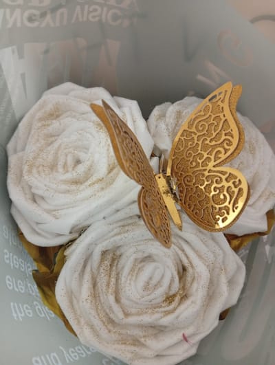 This is a close-up, eye-level, medium shot of a bouquet of white fabric roses, adorned with gold glitter and topped with a metallic gold butterfly decoration. The roses are tightly wound, resembling the petals of real flowers, and are clustered together. The butterfly is positioned on top of the roses, with its wings spread. The background is blurred and appears to be a light gray surface with faint, reflective text, possibly part of packaging or a display. There is no indication of a specific location in Seýdi, Turkmenistan, or any people in the image. The lighting is soft and diffused, highlighting the textures of the roses and the metallic sheen of the butterfly.