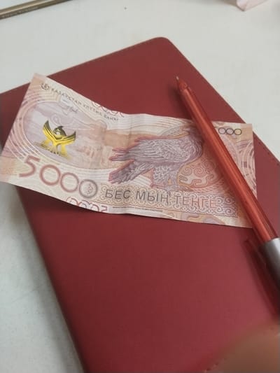 A 5000 Kazakhstani Tenge banknote is placed on a red leather-bound book. A red pen rests diagonally across the banknote and book. The setting appears to be on a white surface, possibly a desk or table. There is no indication that the location is specifically in Töretam, Kazakhstan. No people are present in the image.