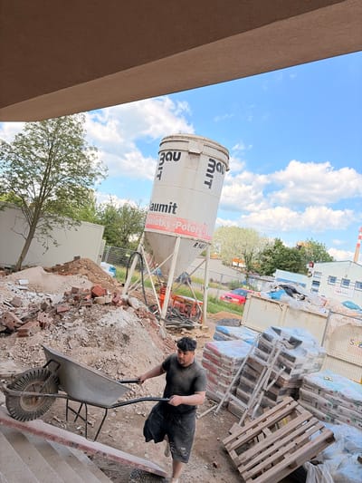This media file captures a construction site scene in Příbram, Czechia, under a bright, cloudy sky, suggesting daytime. The central focus is a man pushing a wheelbarrow up a set of concrete stairs. He is wearing a dark t-shirt and shorts, and appears to be working on the site.

Dominating the background is a large, white cement silo with prominent black lettering that includes "3DU MIT" and "Baumit Omietky-Potery". Beside the silo, stacks of building materials, primarily bags of what appears to be cement or plaster, are wrapped in plastic. These bags display text such as "Baumit" and "PerlaFine". Wooden pallets are also visible, supporting some of the stacked materials.

In the mid-ground, there's a glimpse of a red car, and a chain-link fence separates the construction area from a green, treed background. Further in the distance, a building with multiple windows and a red and white striped smokestack is visible, along with other construction containers. The ground is covered in dirt, rubble, and scattered bricks, typical of an active construction environment. The overall impression is one of ongoing construction work.