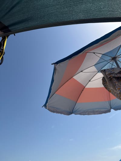 A low-angle shot captures a bright blue sky partially obscured by two beach umbrellas. The umbrella in the foreground, with its canopy made of alternating white, light blue, and coral pink panels, is angled downwards and to the right, with its spokes and a patterned cloth draped over some of them visible. Another darker, teal-colored umbrella is visible in the upper left corner. A yellow strap is attached to the darker umbrella's frame. The scene appears to be set in an outdoor location, likely a beach, under a clear sky, possibly in the city of Gavà, Spain, given the context.