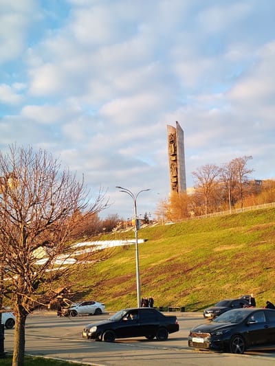 The image shows a street scene in Средний Постол, Russia, featuring the Monument to the Fighters of the Revolution as a prominent landmark. The monument is a tall, concrete structure with sculpted figures. A grassy hill slopes down towards the road. Several cars are parked on the road, including a black sedan, a white sports car with front-end damage, and several other cars. People are standing near the cars and along the sidewalk. A bare tree is visible in the foreground, with its branches silhouetted against the sky. The sky is blue with scattered white clouds. Streetlights line the road.