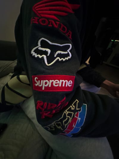 The image displays a close-up view of a person's left arm, adorned with a dark-colored jacket or long-sleeved garment featuring multiple embroidered patches. The perspective is from slightly above and to the side, looking down at the arm.

The most prominent features are the embroidered patches on the sleeve. On the forearm, there is a distinct red rectangular patch with the word "Supreme" embroidered in white block letters. Immediately to the left of the Supreme patch, there is a black patch with a white outline of a fox head logo, representative of Fox Racing. Further to the left, closer to the wearer's wrist area, partially visible red embroidery spells out "HONDA".

Moving towards the upper arm, to the right of the Supreme patch, another Fox Racing logo is visible, this one featuring blue and red elements alongside a gold-outlined fox head. Below this logo, intricate red embroidery is present, parts of which are discernable as "RIPP" or similar stylized text, possibly "R.I.P." or a racing-related acronym.

The fabric of the sleeve appears to be a sturdy, dark material, possibly denim or a similar twill, with visible stitching lines. The person's hand is partially visible at the top, showing a dark wristband or sleeve cuff. To the right of the arm, a portion of a leg, clothed in light grey fabric (possibly sweatpants), is visible, suggesting the person is seated. The background is largely dark and indistinct, indicating low light conditions, possibly indoors at night or in a dimly lit room. No specific activity is clearly depicted other than the person wearing the jacket.