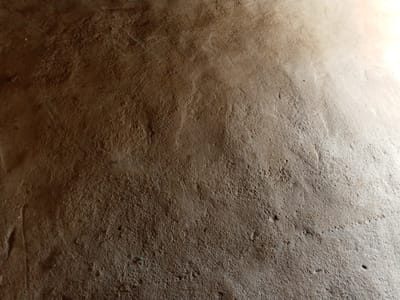 The image depicts a close-up view of a rough, textured surface, likely a wall or ground, with a gradient of light and shadow. The texture is uneven, with small bumps, depressions, and visible striations, suggesting it is made of a material like concrete, mud, or rough plaster. The lighting creates a diagonal effect, with the upper right portion of the image being significantly brighter, indicating a light source from that direction. The color palette is predominantly earthy tones of brown and beige, with the brighter areas appearing lighter, almost white. There are no discernible objects, people, or actions present in the image; it is purely a textural study of a surface. The location in Gindin Dutse, Nigeria, suggests this surface is an element of the local environment, possibly an outdoor wall, pathway, or interior surface of a building.