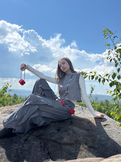 A young female is sitting on a large rock on a sunny day. She is wearing a gray vest over a white long-sleeved shirt and a long gray pleated skirt. She is holding a red cube-shaped pendant on a string in her right hand. The background shows a landscape of green trees and hills under a blue sky with white clouds. The location is Krivel's Mountain in Gubakha, Russia.