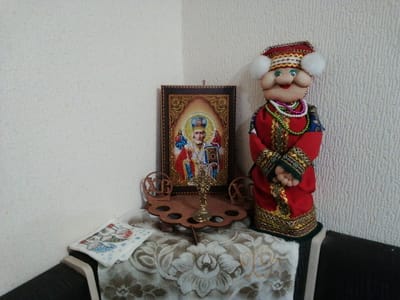 The image displays a still life arrangement on a table covered with a white lace tablecloth. To the left, a small icon of a religious figure, possibly Saint Nicholas, is framed in a dark wooden border adorned with gold beads. In the center, a small wooden stand holds a golden cross and two circular wooden pieces with Cyrillic letters. To the right of the icon, a doll dressed in traditional Russian clothing stands tall. The doll wears a red robe with intricate gold trim, a patterned blue and gold sash, and a red hat with white fluffy pom-poms. It is adorned with several strands of colorful beads around its neck. A small white cloth with a printed pattern of a figure in a blue hat and red coat is placed on the table to the left of the icon. The background consists of a light-colored textured wall. The scene appears to be an indoor display, possibly in a home in посёлок опытного хозяйства «Ермолино», Russia.