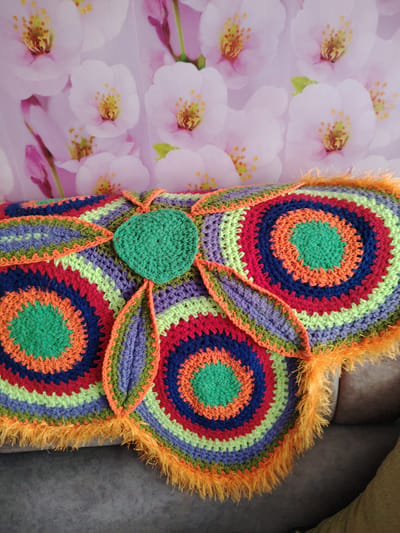 A vibrant and colorful crocheted item, likely a decorative cushion or blanket, rests on a dark gray sofa in an indoor setting. The crocheted piece features a striking design composed of multiple concentric circles, primarily in shades of green, orange, red, dark blue, light green, and purple. A distinct bright green heart-shaped element is centrally placed on the item, with two smaller, leaf-like crocheted extensions branching out from it. The entire perimeter of the crocheted piece is edged with a fluffy, bright orange yarn, adding a soft, textured border.
Behind the crocheted item, a backdrop or wall covering displays a pattern of light pink and white cherry blossoms (sakura) with bright yellow stamens, creating a soft, floral aesthetic. The lighting suggests daytime, enhancing the vividness of the colors. No people are visible, nor is any text present in the image. The scene conveys a sense of handmade warmth and cheerful decor. This image was taken in Yanaul, Russia.
