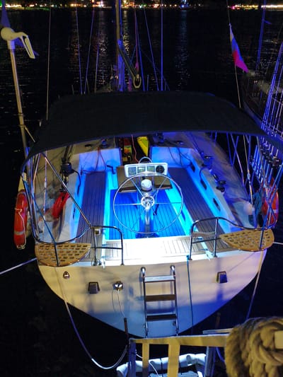 An elevated, direct overhead shot captures the rear deck of a white sailboat at night in the city of Krinitsa, Russia. The deck is illuminated with bright blue lights, creating a striking contrast with the dark water surrounding the vessel. A black canopy covers the central portion of the deck, and a silver steering wheel is prominently displayed. To the left, a red life preserver hangs from the railing, and a coiled red rope is visible below it. Various ropes and rigging are also apparent on the deck and extending upwards. A silver ladder is positioned on the stern of the boat. In the background, the lights of buildings and other vessels are visible along the shoreline, reflecting on the water. The scene suggests a peaceful, illuminated nighttime setting on the water.