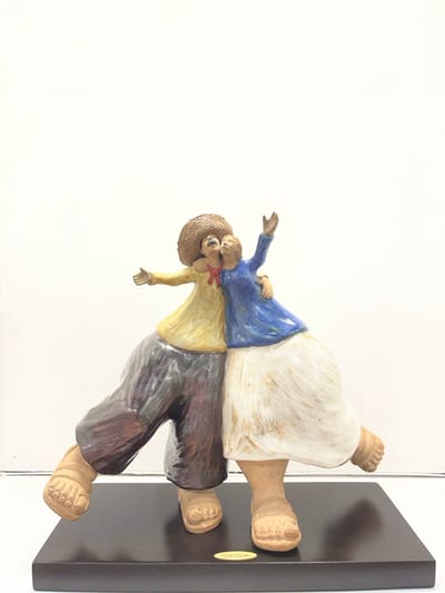 A ceramic sculpture depicts a person giving another a piggyback ride. The bottom figure wears a yellow shirt, brown pants, and a hat, while the upper figure is dressed in a blue shirt and white skirt. The figures have exaggerated feet and are attached to a black rectangular stand. The sculpture is positioned against a white background.
