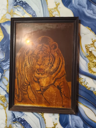 A rectangular framed artwork is centered on a patterned wall in Cileungsi, Indonesia. The frame is dark brown or black with a beveled profile. The artwork encased within depicts a tiger, facing forward and slightly to the viewer's left, rendered in warm orange, brown, and dark tones. The tiger's mouth is open, revealing teeth, and its body displays prominent dark stripes. A bright, circular reflection or glare obscures the upper-left central portion of the artwork's protective covering, partially obscuring the tiger's chest and lower face. A fainter reflection is present near the lower-right edge. The background wall features an abstract, organic pattern composed of swirling shapes in shades of blue and white, interspersed with textured, granular clusters of metallic gold.