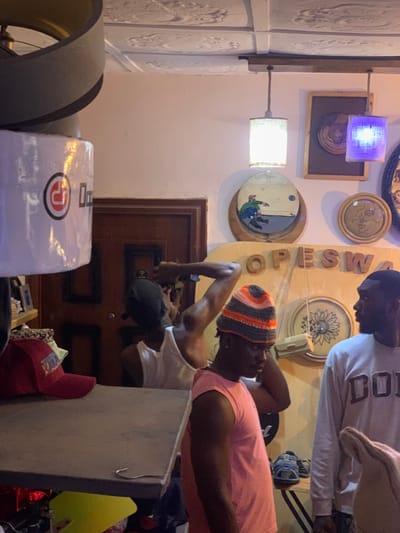 The image shows the interior of a shop, with the address being Ita Oniyan, Nigeria (latitude: 7.282, longitude: 5.161). There are three men in the shop, one of whom is wearing a white tank top and is facing away from the camera. The other two men are facing the camera, and one is wearing a pink tank top and a multi-colored beanie hat. The man in the beanie is touching his hat with his right hand. The other man is wearing a gray sweatshirt with the word "DOU" on it. The shop is decorated with wooden wall art, including a circular framed picture with a man in it, a sunflower-shaped piece of art, and the word "OPESWA" in wooden letters. The shop also has two pendant lights, one of which is on, and a number of hats and other objects sitting on a shelf. There is a closed wooden door to the left side of the image. There are sandals on a shelf in the foreground of the picture. There is a large, open ceiling fan in the corner of the image, with a gray metal covering around it. The image is lit by overhead lights and the daylight that is coming through the doorway in the background.