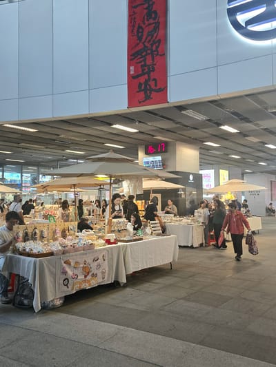 The image captures an outdoor market scene with several stalls set up under white umbrellas. The stalls display various merchandise, including trinkets, jewelry, and small decorative items. People are browsing and shopping at the stalls, with some interacting with vendors. The overall environment appears to be a pedestrian walkway or plaza, with a large building structure overhead. A digital clock displays "16:17". A large red banner with Chinese calligraphy hangs from the overhead structure. In the background, a circular logo is visible on the building's facade. This appears to be a street market, possibly in a public area or transportation hub, within Kaohsiung, Taiwan, given the cultural context of the calligraphy and the architecture.