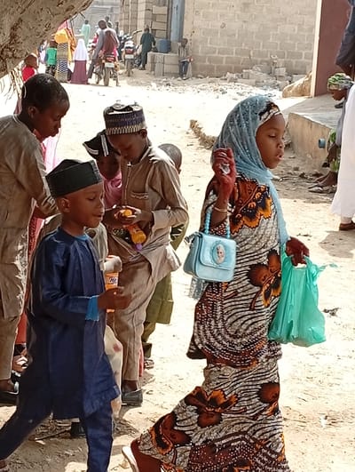This medium shot captures a lively street scene in Fanisau, Nigeria, bathed in bright daylight. The foreground is dominated by several children, with more people and buildings visible in the dusty background.
On the right, a young girl walks towards the right side of the frame. She is dressed in a long, flowing garment with a distinctive brown and orange floral pattern over a lighter background, paired with a light blue hijab. She carries a small, light blue shoulder bag adorned with a "Frozen" character, and a translucent green plastic bag in her right hand. Her left hand is decorated with intricate henna patterns and she holds a small, light-colored item.
To her left, a young boy walks in a dark blue traditional outfit, including a top and trousers, and a dark green cap. He holds an orange-labeled bottle, possibly a drink, in his right hand. Another boy, wearing a tan traditional shirt and a patterned cap, is positioned slightly behind him, looking down and holding an orange object, likely a fruit or snack. On the far left, another child in a light-colored top is partially visible.
The background reveals a dirt path or road with simple, light-colored buildings, some appearing to be constructed from cinder blocks. Several adults and other children are scattered throughout the scene; some are walking, others are stationary, and a person on a motorcycle is visible further back. A large tree trunk provides shade on the far left. The overall impression is one of everyday activity in a warm, semi-urban or rural setting. The "Frozen" branding on the girl's bag and possibly "HP" on the boy's bottle are the only discernible texts.