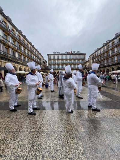 A group of people dressed in white chef's uniforms and hats are marching in a line on a wet cobbled street in the Plaza de la Constitución, San Sebastian, Spain (latitude: 43.324, longitude: -1.985). The group is holding drumsticks and hitting small wooden barrels. Several other people in various costumes are scattered across the square in the background. The sky is overcast and it appears to be raining. Buildings with balconies line the street on both sides of the square.