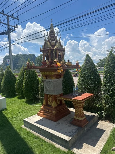 In Tambon Phraek Ha, Thailand, a roadside shrine stands prominently in the foreground. The shrine, made of wood with intricate golden carvings, is adorned with a grey cloth wrapped around its base and a yellow flower garland. To its right, a small wooden table holds two plastic bottles of water. The shrine is situated on a concrete platform surrounded by a lush green lawn and neatly trimmed cypress trees. In the background, power lines crisscross the bright blue sky, dotted with fluffy white clouds. Beyond the trees, a road is visible, with a motorbike passing by, and a gas station with a blue and red logo further in the distance. A large, verdant mountain range forms the backdrop. The scene is bathed in bright daylight, casting distinct shadows.