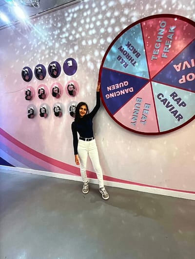 The image shows a woman lying on her back with her arms outstretched, appearing to be resting on a wall or floor. She is wearing a dark blue long-sleeved shirt, white pants, and white sneakers. Her legs are extended towards the right side of the frame, and her arms are stretched upwards and to the sides.
Behind her, a large, circular, colorful spinning wheel is visible, positioned vertically. The wheel is divided into several segments, each with text printed on it. The visible text includes "OP IVA", "RAP CAVIAR", "BEAT BURRY", "DANCING QUEEN", "R&B THROW BACK", and "TECHNO FREAK". The colors of the segments are pink, blue, and light blue with white text.
To the left of the woman and below the spinning wheel, there is a grid of circular objects. These appear to be reflective buttons or lights, arranged in rows. Some are purple, and some are pink, with a darker circular element inside. The background behind the woman and the wheel is a textured wall with a striped pattern, featuring pink, white, and gray stripes. The overall lighting suggests an indoor setting, possibly a studio or entertainment venue. The woman has a slight smile on her face, giving an impression of relaxation or fun.