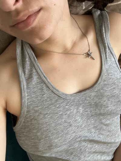 The image shows a close-up view of a person's upper torso and neck, with a focus on their décolletage and chin area. The person is wearing a grey tank top and a silver necklace with a cross pendant. The background is indistinct, but appears to be a couch or bed with a plaid blanket. The lighting is soft and natural, creating a relaxed and intimate atmosphere. There is no indication of the image being taken in Postoyata, Ukraine.