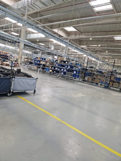 The image depicts the interior of a large, modern warehouse facility. The scene is dominated by a vast, industrial space with high ceilings supported by metal beams. Natural light streams in through skylights, illuminating the grey concrete floor. Racks and shelves are visible in the background, filled with various boxes and blue containers, suggesting storage and organization of goods. In the foreground, two grey fabric carts with metal frames and wheels are positioned. The cart on the left is partially obscured by the one on the right, which is filled with black plastic bags. A prominent yellow line is painted on the floor, running diagonally across the image, likely indicating a pathway or safety zone. There are no people visible in the image. The overall environment suggests a functional and operational warehouse. The location of Bahovitsa, Bulgaria is not visually represented in the image itself.