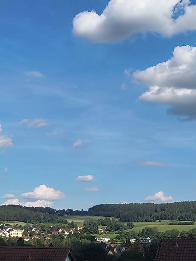 The image depicts a bright, sunny day in Oberviechtach, Germany. The scene is dominated by a vast expanse of blue sky filled with fluffy white cumulus clouds. In the distance, a verdant landscape unfolds, characterized by rolling green fields and a dense, dark green forest that lines the horizon. Nestled within the trees and fields are a scattering of houses with red-tiled roofs, suggesting a rural or semi-rural residential area.
The image is taken from an elevated perspective, possibly from a rooftop or a high window, as a section of a dark red tiled roof is visible in the foreground, cropped along the right edge. The sun is high in the sky, casting bright light and creating distinct shadows, indicating it is likely midday or early afternoon. The weather appears to be fair and warm, typical of a summer day.
There are no people or animals clearly visible in the frame. A small, dark speck that could be a bird or an insect is present in the sky towards the left of the image. The overall mood conveyed by the image is one of peace and tranquility, showcasing the beauty of the German countryside on a clear day. No text is visible in the image.