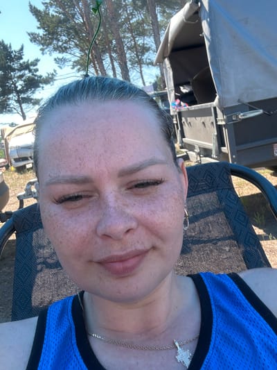 A woman with grey hair pulled back and a small green sprout attached to the top of her head is sitting in a black mesh chair outdoors. She is wearing a blue tank top with black trim and a silver chain with a cross pendant. The background shows trees, a trailer, and a light-colored vehicle. The scene appears to be a sunny day in a rural or campsite setting.