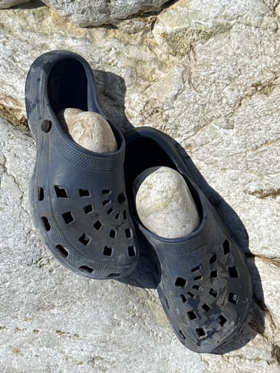 Two dark blue Croc-style shoes are placed on a rough, light-colored rocky surface. Each shoe contains a light-colored, smooth, rounded stone. The shoes are positioned side-by-side, with the left shoe slightly overlapping the right. The rocky surface appears to be outdoors, illuminated by sunlight, creating shadows around the shoes. The environment is consistent with a natural, possibly coastal or mountainous area. The image does not contain any information about the city of Donji Katun, Montenegro, nor does it depict any people or specific actions.