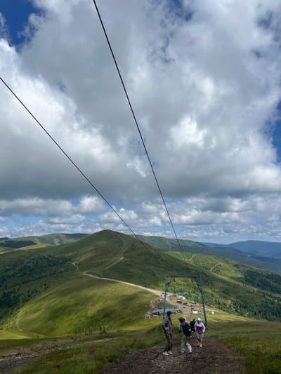 The image shows a cable car station in a mountainous landscape, likely in Chorna Tysa, Ukraine. In the foreground, three young adults stand on a dirt path. One person on the left wears a plaid shirt and dark pants. Another person in the center wears a backpack and light-colored pants. A third person on the right wears a white hoodie and blue shorts.
Behind them, a blue cable car support structure is visible, with cables stretching upwards towards the sky. In the mid-ground, there are several buildings and vehicles at the cable car station. The background consists of rolling green mountains covered in grass and trees, with winding dirt roads visible on the slopes. The sky is partly cloudy with patches of blue. The scene appears to be a tourist or recreational area in a natural mountain setting.