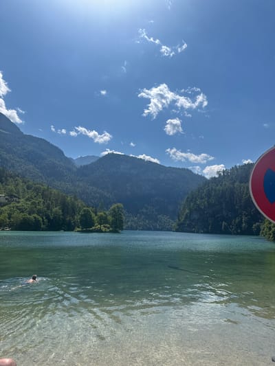 This is an eye-level, medium shot of Königssee lake in Schönau am Königssee, Germany. The sun is shining brightly, casting light across the calm, clear water, which reveals the rocky lakebed in the foreground. A person is swimming in the lake, facing away from the camera. The background features a densely forested mountain range that rises steeply from the lake's edge. A small island with a single tree is visible in the middle distance. On the right side of the frame, a red and blue "no parking" sign is partially visible. The sky is a clear blue with scattered white clouds.