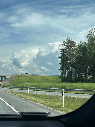 The image depicts a roadside view on a cloudy day, likely from inside a vehicle, in Saku, Estonia. In the foreground, the asphalt road surface is visible with white lane markings. A metal guardrail runs along the side of the road, with white posts with black rectangular markers. Beyond the guardrail is a grassy embankment. In the middle ground, a raised road or bridge is visible with a dark-colored car driving on it. The background features a line of green trees and a large, dramatic cloud formation. The sky is blue with scattered clouds, including some darker, more voluminous clouds that suggest potential rain. The overall impression is of a typical highway scene in a rural or suburban area.