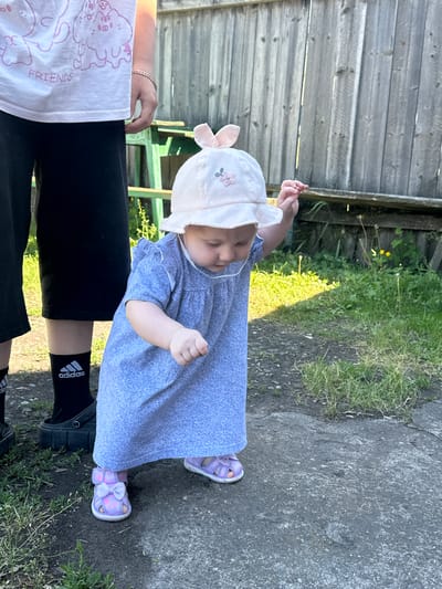 The image shows a young child in a blue dress and a pink hat with bunny ears, standing on a dirt and concrete path outdoors. The child is facing forward and slightly to the right, with their right arm extended. To the left of the child, a person's legs are visible, wearing black shorts and black socks with the Adidas logo. Behind the child is a wooden fence and some greenery, including grass and a few small yellow flowers. The scene appears to be in a residential area. There are no clear indicators of the specific location within Novoyeniseyka, Russia, beyond being an outdoor setting.