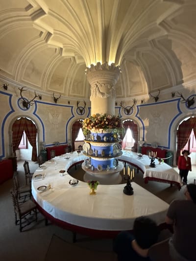 The image shows a large circular room in what appears to be a palace or castle. The room is located in the city of Sintra, Portugal. A large round table dominates the center of the room, covered with a white tablecloth and set with plates. In the center of the table is a large, tiered centerpiece adorned with flowers and plates. A large decorative column rises from the center of the room to the ceiling. The ceiling is high and vaulted, with ornate architectural details.
Along the walls are arched windows draped with red curtains. There are deer antlers mounted on the walls. Several people are in the room, including a person in a red jacket, seated people, and other blurred figures. The room has a formal, historic atmosphere.