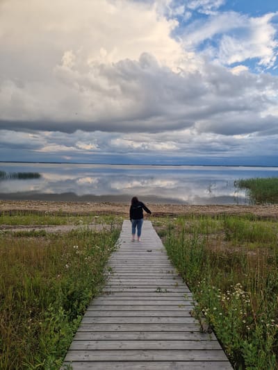 A person stands on a wooden boardwalk, facing a calm body of water. The boardwalk is made of weathered gray planks and runs diagonally across the right side of the frame, bordered by tall green grass and some wildflowers. The water reflects the dramatic sky, which is filled with a mix of bright white and dark gray clouds, suggesting an overcast or rapidly changing weather day. The horizon line is clearly visible where the water meets the sky. The person is wearing a dark jacket and light-colored pants. Their pose, with one arm slightly raised, suggests contemplation or enjoyment of the scenery. The overall impression is one of peace and connection with nature. The time of day appears to be late afternoon or early evening, given the quality of light. The location is identified as Īdeņa, Latvia. There is no visible text in the image.