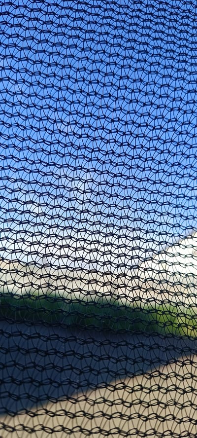 The image is a close-up shot of a black net with a blue sky and a hint of green landscape visible through it. The net creates a repetitive pattern of interconnected loops, obscuring the view of the background. The sky is clear and bright, suggesting it is daytime with good weather. The lower portion of the image shows a blurred area of green, likely grass or trees, and a lighter, possibly sandy or paved, surface. Shadows are cast by the net onto the ground below, adding depth to the scene. There is no discernible text or activity taking place. The overall impression is abstract due to the dominant pattern of the net.