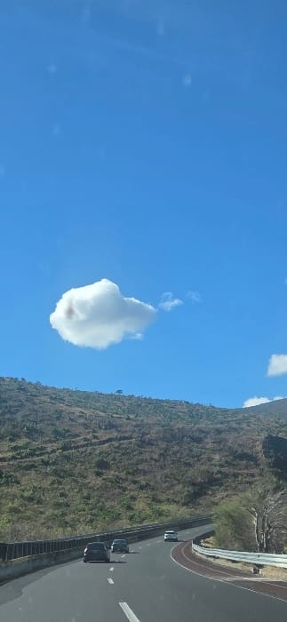 A cloud resembling a finger pointing upward dominates the blue sky. Adjacent to the sky, a mountainside covered in dense, textured greenery slopes down. A highway with moving vehicles is visible next to the mountainside. The image is rotated sideways.