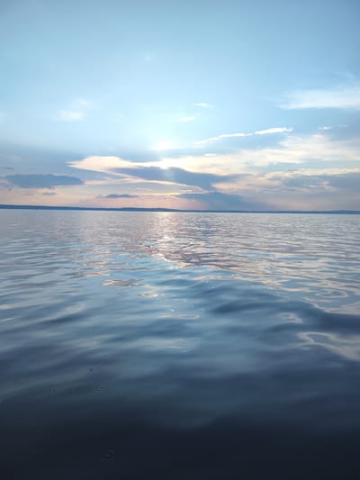 The image depicts a serene body of water, likely a lake or a large river, under a partly cloudy sky. The sky is a pale blue with scattered white and gray clouds, some of which are illuminated by the setting sun, casting rays of light that are visible through the clouds. The sun itself is not directly visible but its presence is indicated by the bright reflection on the water's surface. The water is calm with gentle ripples, reflecting the colors of the sky and clouds. The horizon line is visible, separating the sky from the water, and a faint, dark landmass can be seen in the distance. The foreground of the image shows the water in more detail, with the ripples creating patterns of light and shadow. Several small, circular disturbances are visible on the dark surface of the water in the lower part of the image, possibly from raindrops or small creatures. There are no people, boats, or structures visible in the image. The scene appears to be in a natural, undeveloped area, and the location in the city of Галево, Russia, suggests a possibly tranquil, outdoor setting near a body of water within or adjacent to the city.