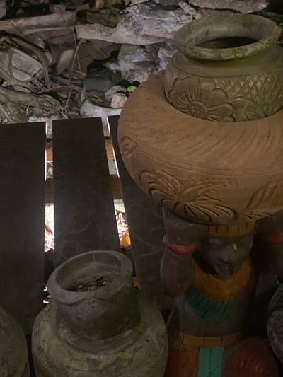 The scene is outdoors, likely in a garden or outdoor display area in Baan Don Sai, Thailand. The focal point is a collection of large, ornate terracotta pots. One prominent pot is light brown with carved floral patterns around the top. Another pot, grey and weathered, shows its interior filled with pebbles or dirt. These pots rest on dark wooden slats or a platform, with visible space between the slats revealing leaf litter and debris underneath. The background shows rocks, roots, and other ground covering materials. A portion of a decorative statue is also visible, featuring painted sections in yellow, green, and red. The lighting appears dim, perhaps during the evening or under heavy shade.