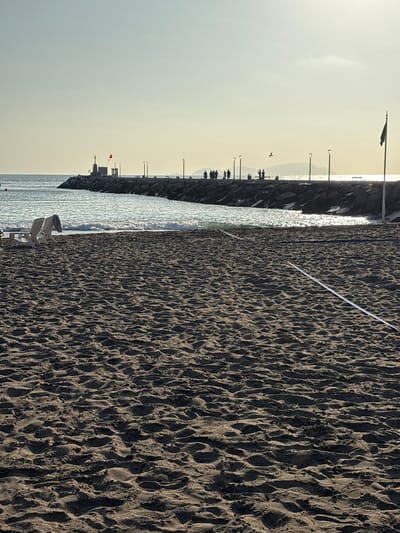 The image depicts a beach scene in Chorrillos, Peru, likely during a late afternoon or early morning given the low angle of the sun and the soft lighting. In the foreground, dark sand with subtle ripples and indentations covers the entire lower half of the frame, suggesting it might be a volleyball court due to a white line marker visible diagonally across the sand. Two empty white lounge chairs with a towel draped over one are positioned near the water's edge, hinting at a relaxed atmosphere.
The middle ground features the gentle surf of the ocean meeting a rocky breakwater that extends into the sea. Along the breakwater, several lampposts are visible, and a small structure with a red flag atop it stands near the shore. Silhouettes of people are scattered along the breakwater, some appearing to walk, others possibly observing the scenery. A flag with a dark emblem is mounted on a flagpole at the far end of the breakwater.
In the background, the ocean stretches towards the horizon, where a hazy outline of distant landmasses can be discerned against a pale sky. The overall ambiance is serene, capturing a quiet moment at the Chorrillos coastline.