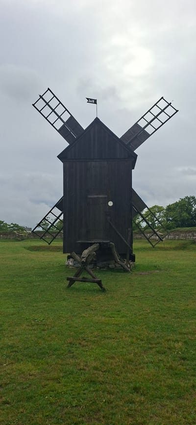 A dark wooden windmill with four sails, two of which are partially visible, stands in a grassy field under a cloudy sky. A black flag with "2018" printed in white is attached to the top of the windmill. In the foreground, a wooden structure, possibly a prop or a part of the windmill's mechanism, is visible on the grass. The background shows a low stone wall and trees. The image appears to be taken in Kuressaare, Estonia, given the context of similar windmills found in the region. There are no people or animals visible in the image. The overall scene is serene and historical, with a focus on the rustic architecture of the windmill.