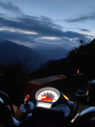 The image is a first-person view from a motorcycle dashboard at dusk in Siumsa, India. The dashboard's illuminated speedometer, showing a red needle, is in the foreground. The background features a dramatic mountain landscape under a cloudy sky, with the faint outline of a crescent moon visible. The scene is dimly lit, suggesting the transition from day to night. Trees and a road marker are also present in the background.