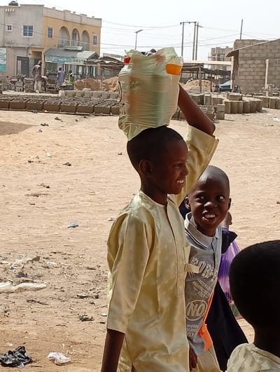 This image, taken in Fanisau, Nigeria, depicts a bright, sunny outdoor scene featuring several young boys. In the foreground, a boy, approximately 8-10 years old, smiles widely as he carries a clear plastic bag, seemingly filled with bottled drinks, balanced on his head. He is dressed in a light yellow, long-sleeved tunic. Beside him, another boy of similar age grins towards the camera, wearing a grey long-sleeved shirt with partially visible English text, including "ANDREW" and "NO". Other children are partially visible in the immediate background.
The ground is unpaved, dusty, and strewn with scattered litter and debris. In the mid-ground, numerous grey rectangular blocks, likely concrete or mud bricks, are neatly arranged, indicating a construction material site or an area for brick production. The background features various buildings under a clear sky. On the left, a two-story building with arched windows stands next to a single-story structure with a green sign reading "SABO". On the right, uncompleted structures made of cinder blocks are visible, along with overhead power lines and poles. The strong sunlight suggests it is midday or early afternoon. The children's expressions convey happiness and amusement.