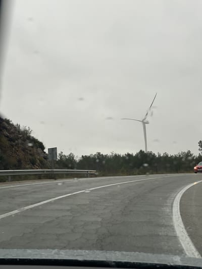 A roadside view shows a wind turbine standing tall against a gray, overcast sky. The road, wet from rain, curves into the distance with a guardrail visible. A line of trees separates the road from the landscape where the wind turbine is situated. The scene suggests a rural or industrial landscape on a dreary day.