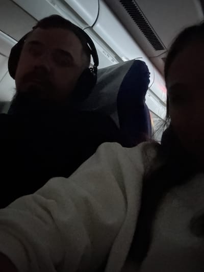 The image depicts two people on what appears to be an airplane. One person is wearing headphones. The location is indicated by the mention of Stolbishche, Russia; however, the image itself does not provide visual confirmation of this specific city. The environment shows an airplane interior, including a window and overhead compartments.