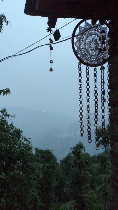 The image, taken from a low angle in Qila Nathu Singh, India, features a dreamcatcher and strings of metal chain hanging in the foreground against a backdrop of an overcast sky. The dreamcatcher is circular with an intricate web design and several small objects attached. Adjacent to the dreamcatcher, multiple strands of metal chain, composed of interconnected rings, are suspended horizontally. To the right, a dense cluster of trees and foliage obscures the view, creating a dark contrast against the bright sky. The sky is uniformly light with three distant points of light. A few thin strings with small objects attached hang from the left. The objects are small and do not display many details. The lighting is subdued, indicating either twilight or overcast conditions.